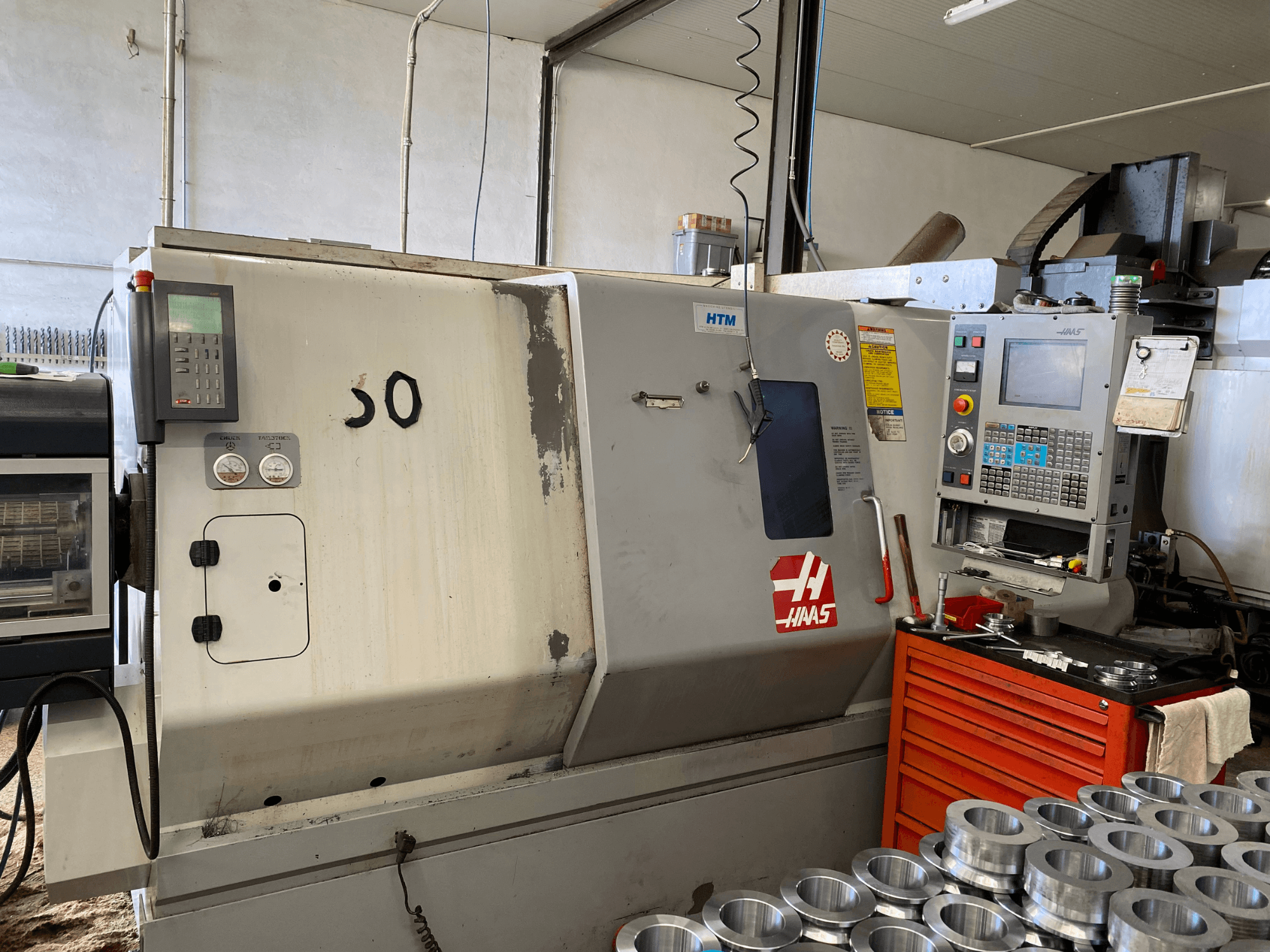 Haas CNC machine with control panel, front view, showing operation buttons, gauges, and metal parts stacked nearby.