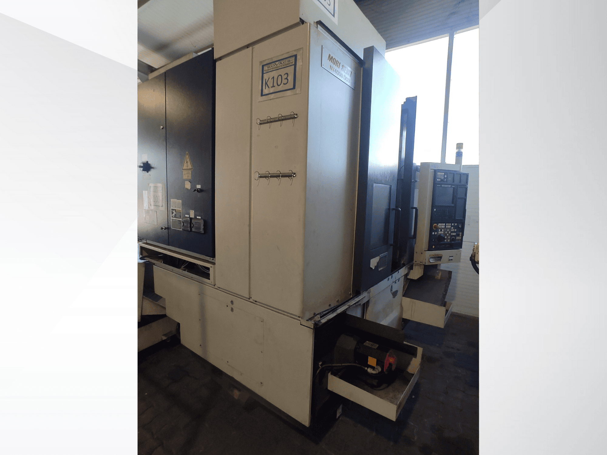 Mori Seiki K103 CNC machine in front view, featuring control panel, safety labels, and electrical components visible.