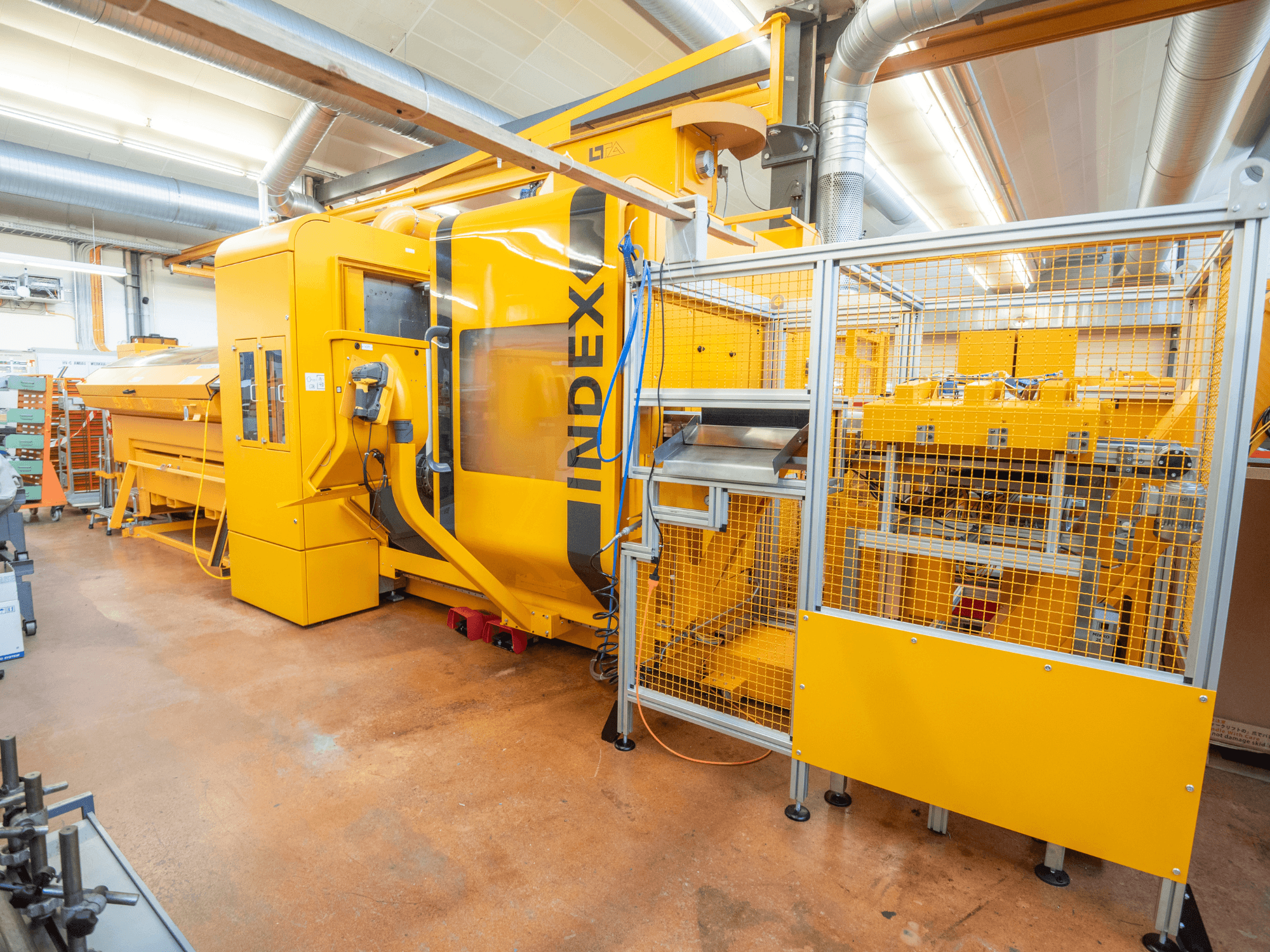 Yellow INDEX machine in a workshop, front view. Features safety enclosure, control panel, and multiple operational components.