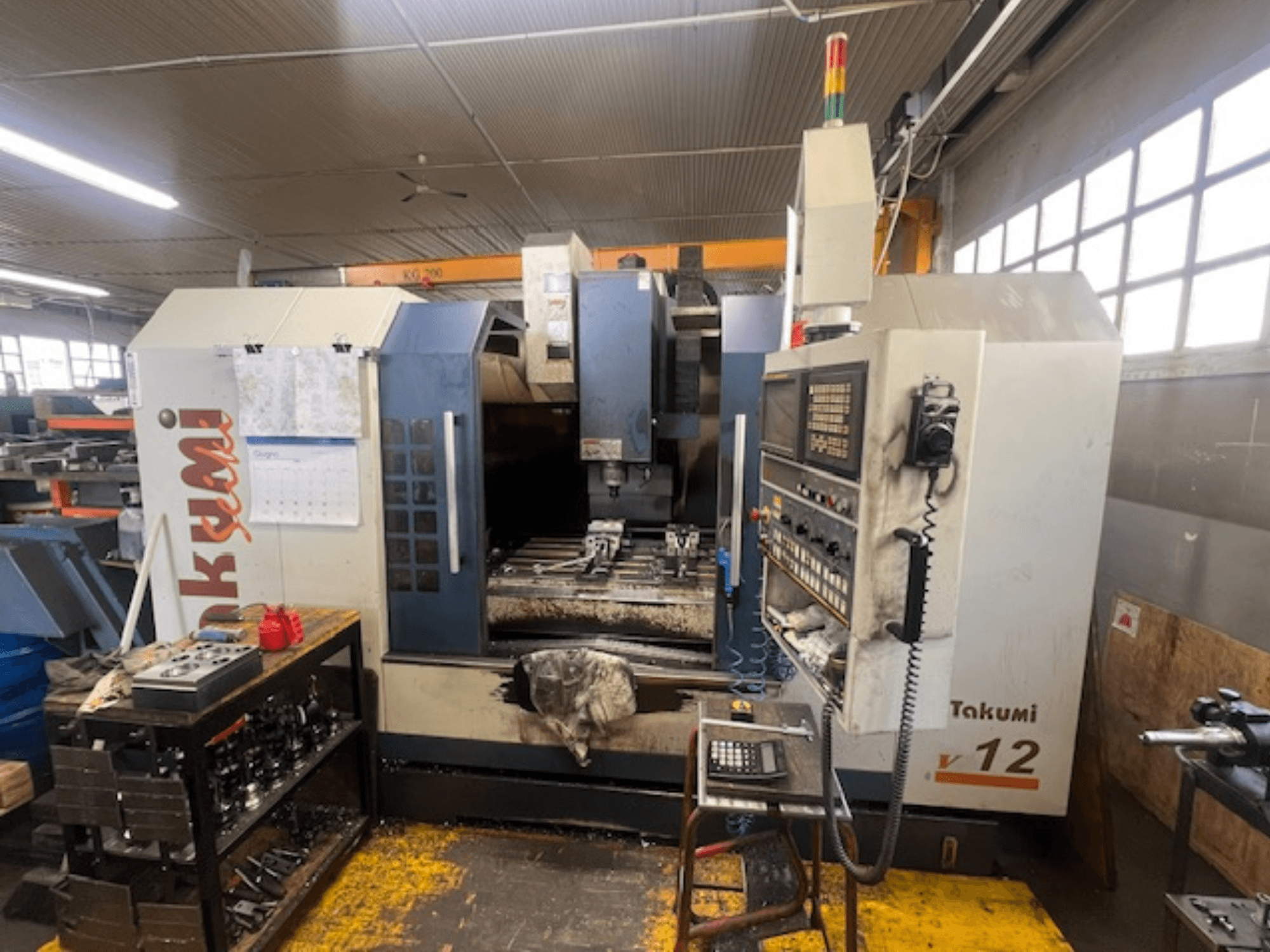 Takumi V12 CNC milling machine in side view, showing control panel, cutting area, and tools on the workstation.