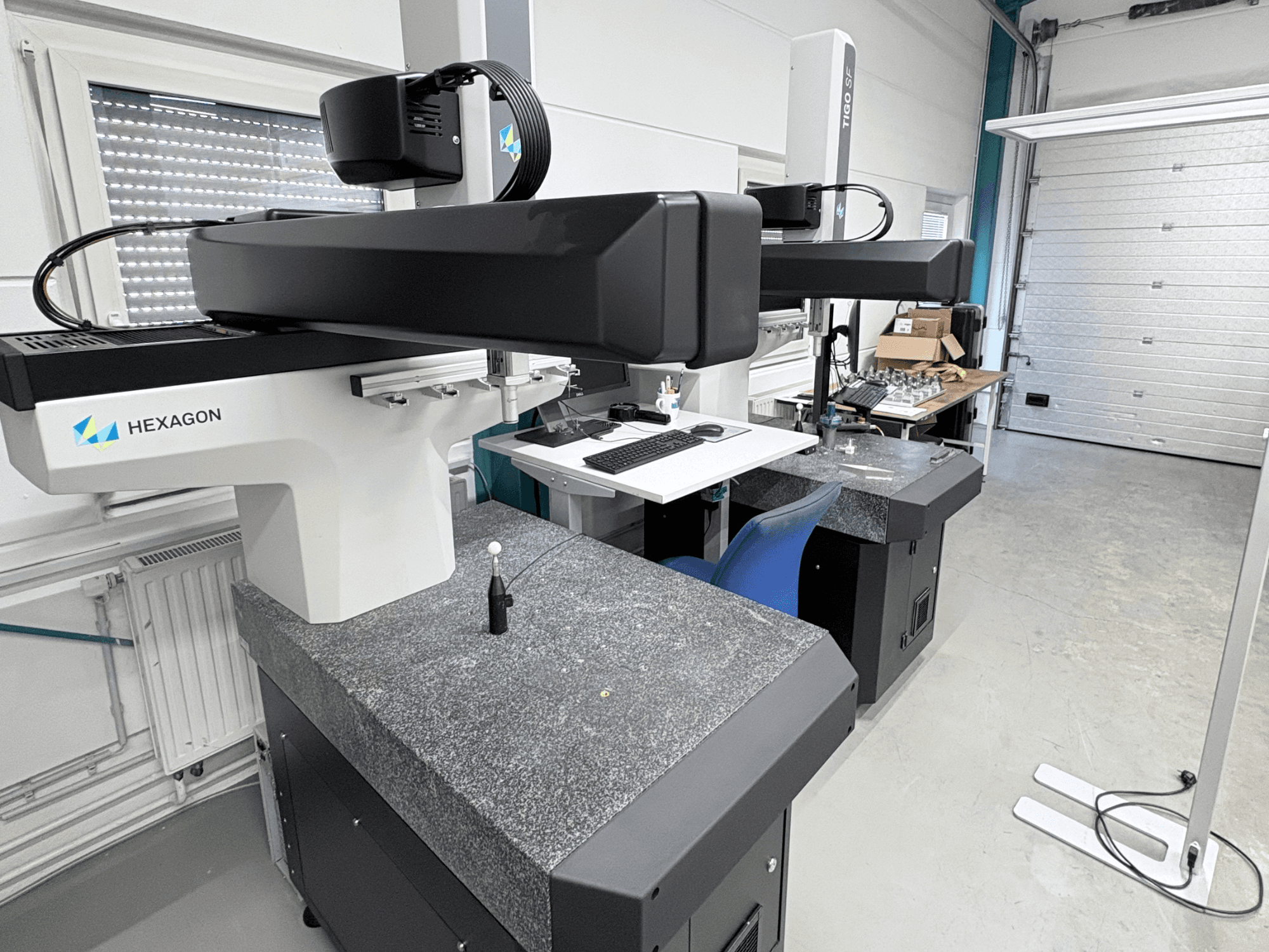 Hexagon Tigo SF CMM in workshop, featuring a granite base, measurement probe, and nearby workstation with tools.