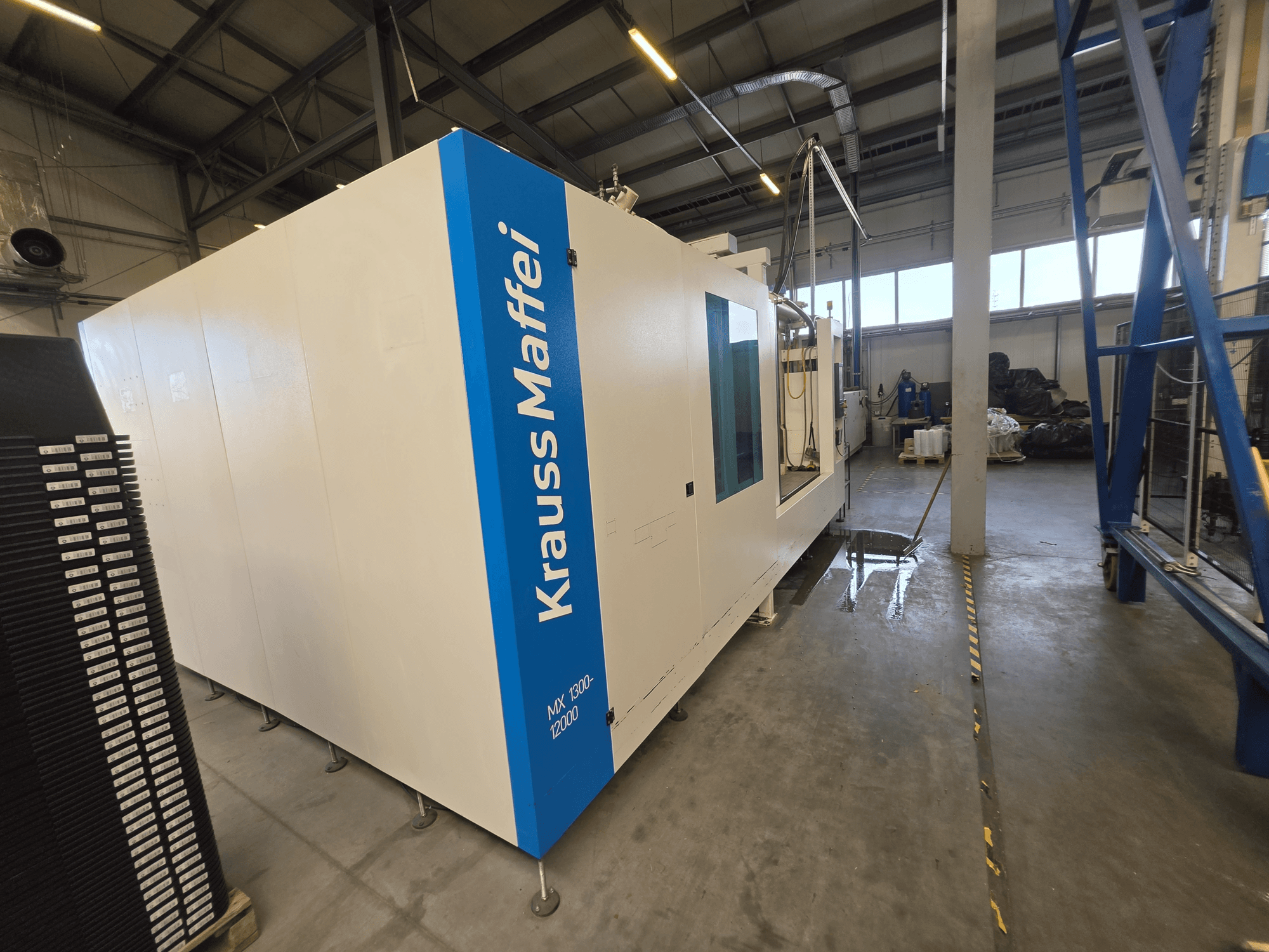Krauss Maffei MX 1300-12000 hydraulic injection molding machine in side view, showcasing its large white chassis and blue branding.