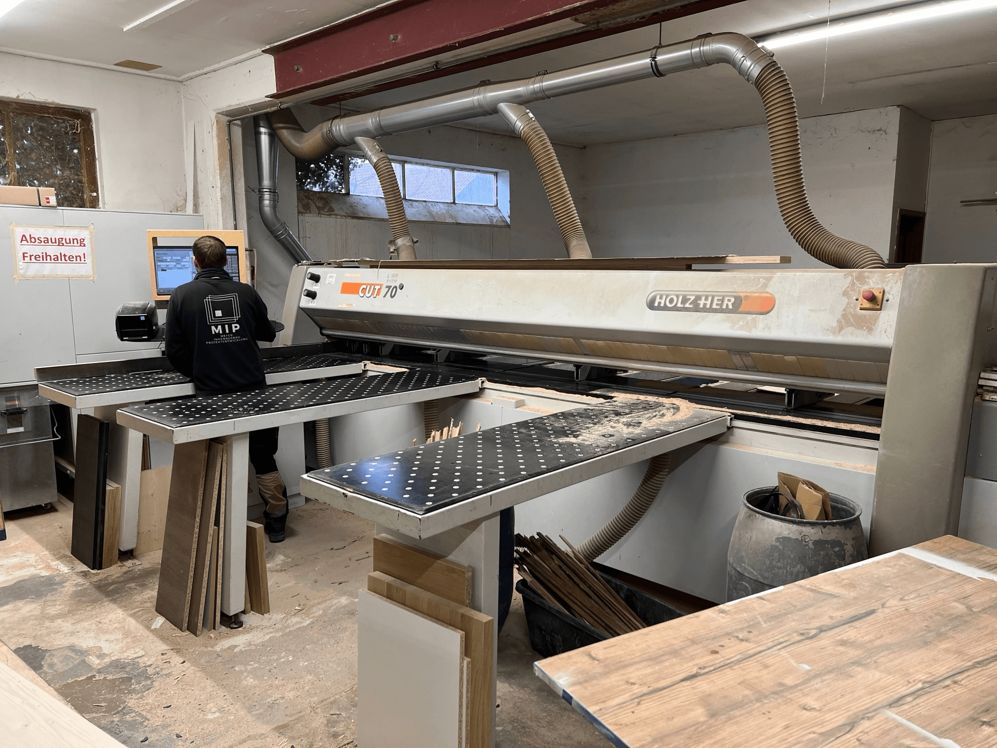Holzher CUT 70 saw in a workshop, shown from the side, with a worker operating controls and wood panels on the tables.