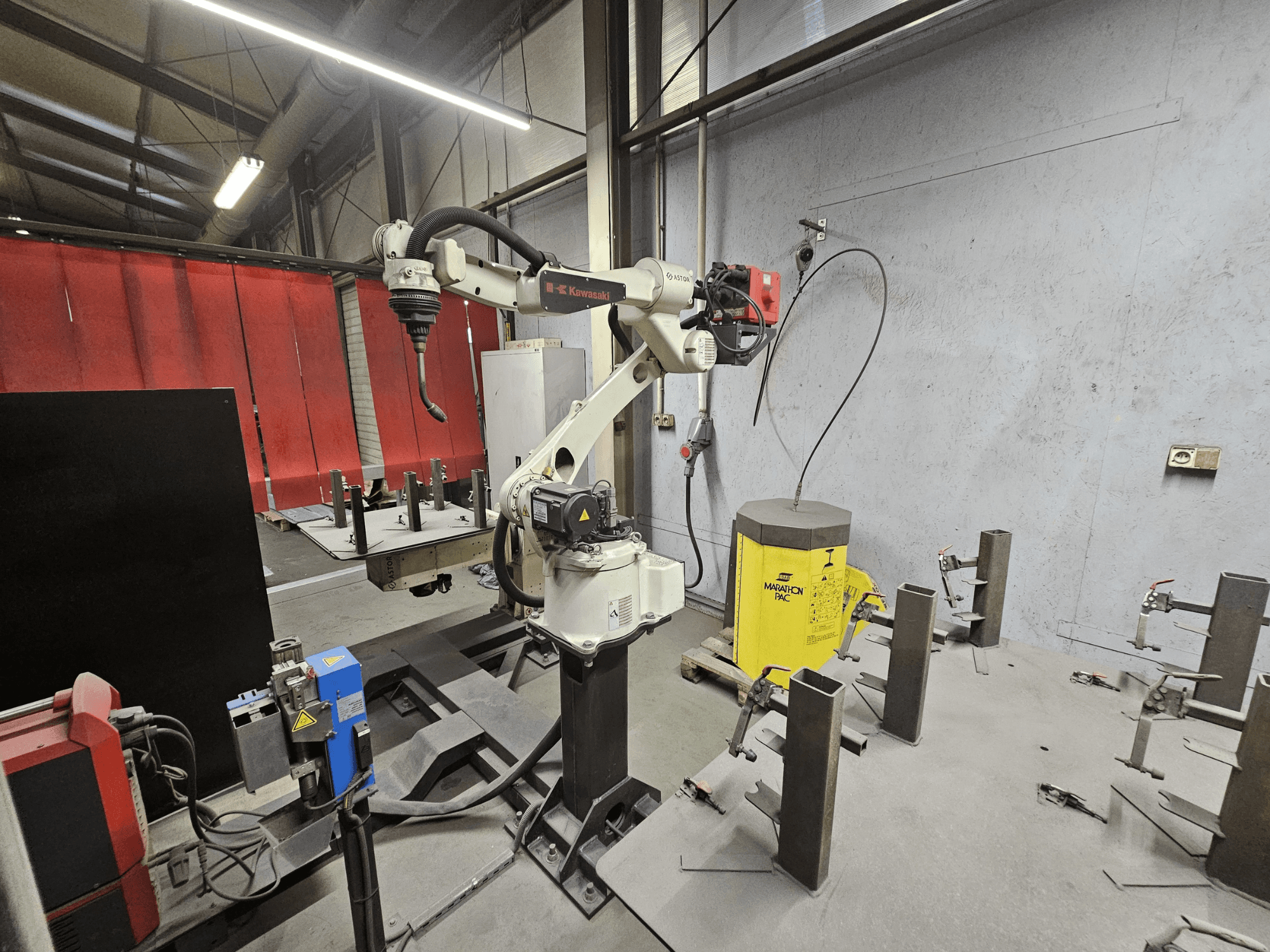 Kawasaki robotic arm in a factory, side view, with welding equipment and metal parts on a work table, industrial setting.