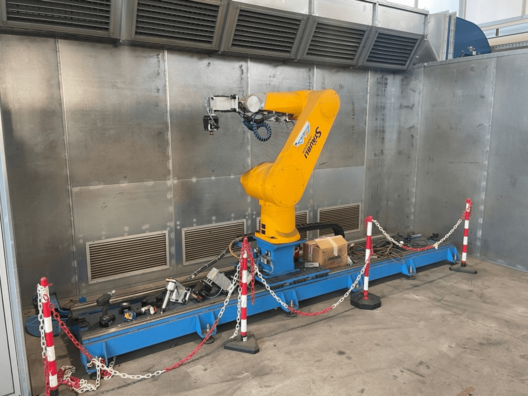 STAUBLI CS8C-RX160 robot arm in a workshop, side view, with cables and various tools on a blue base surrounded by protective barriers.