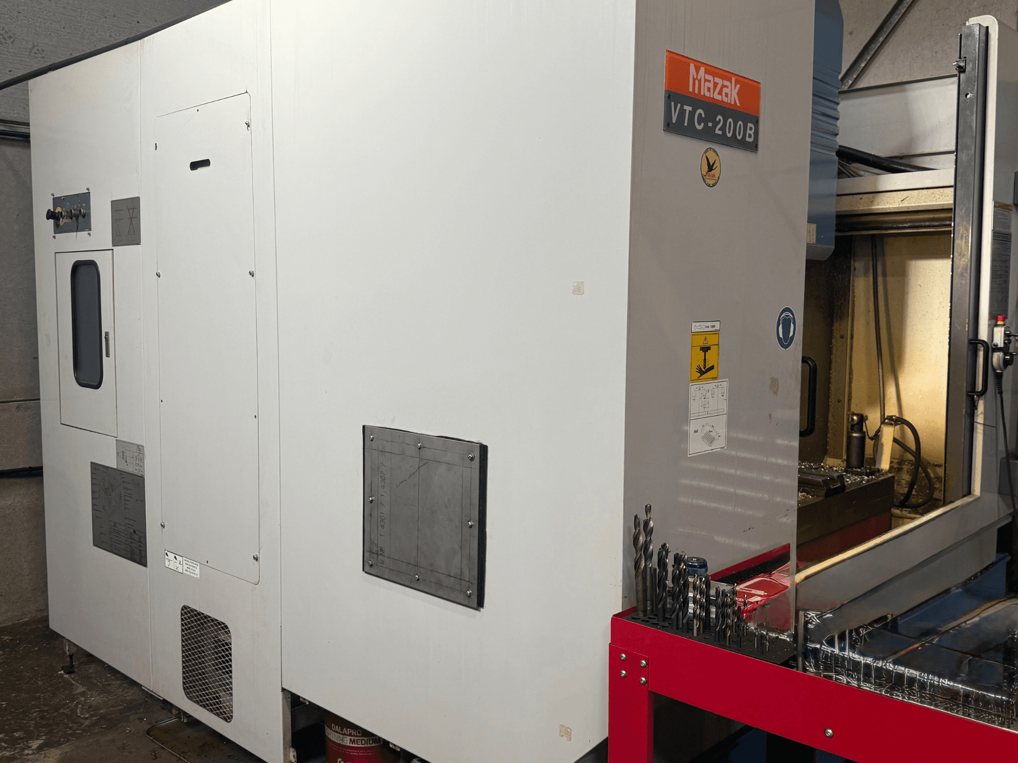 Mazak VTC-200B vertical machining center in side view, featuring labeled controls and drill bits on red storage rack.