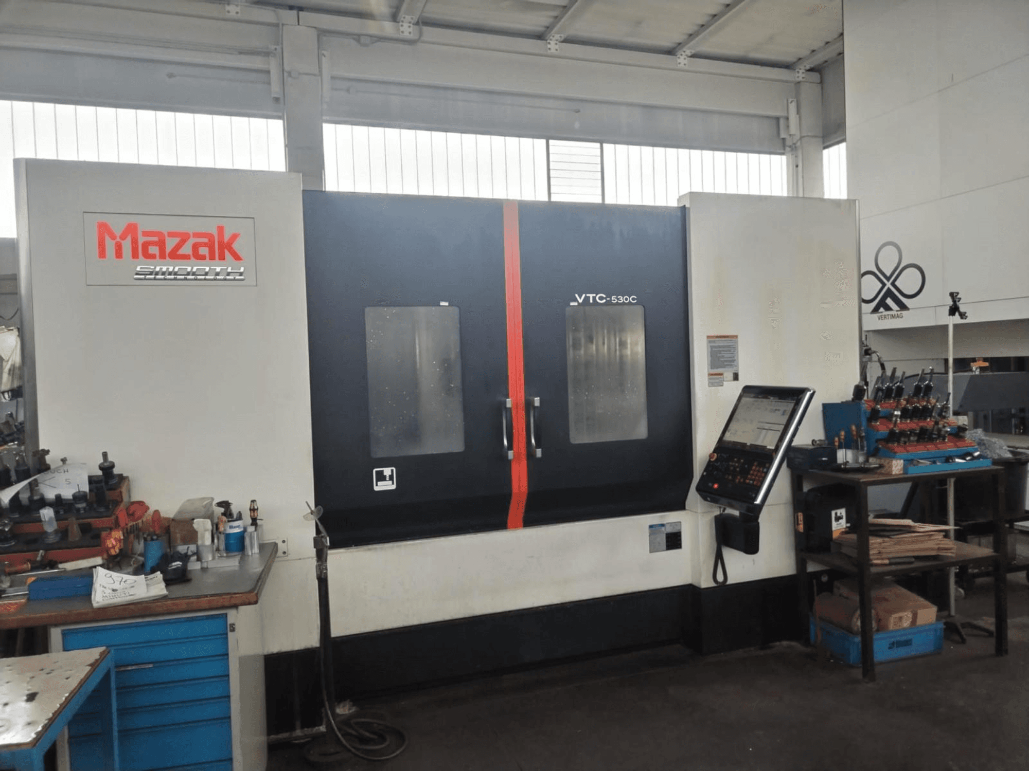 Mazak VTC-530C vertical machining center in a workshop, featuring a control panel and tools on a side table.