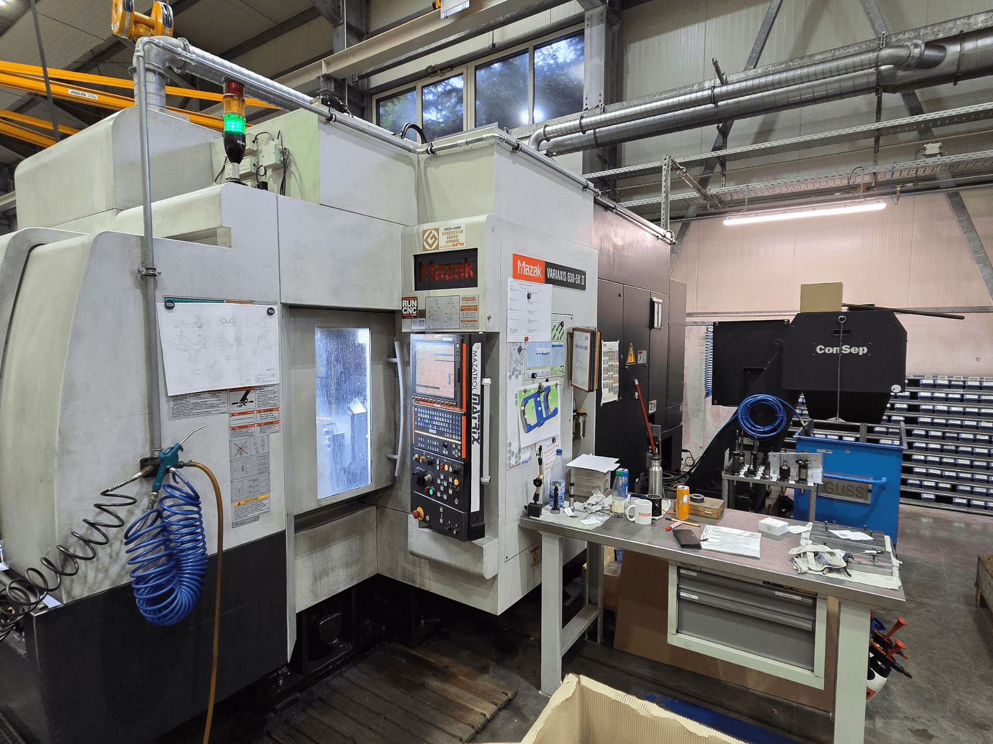 Mazak Variaxis 630-5X CNC machine in a workshop, front view with control panel, hose, and accessories on side table.