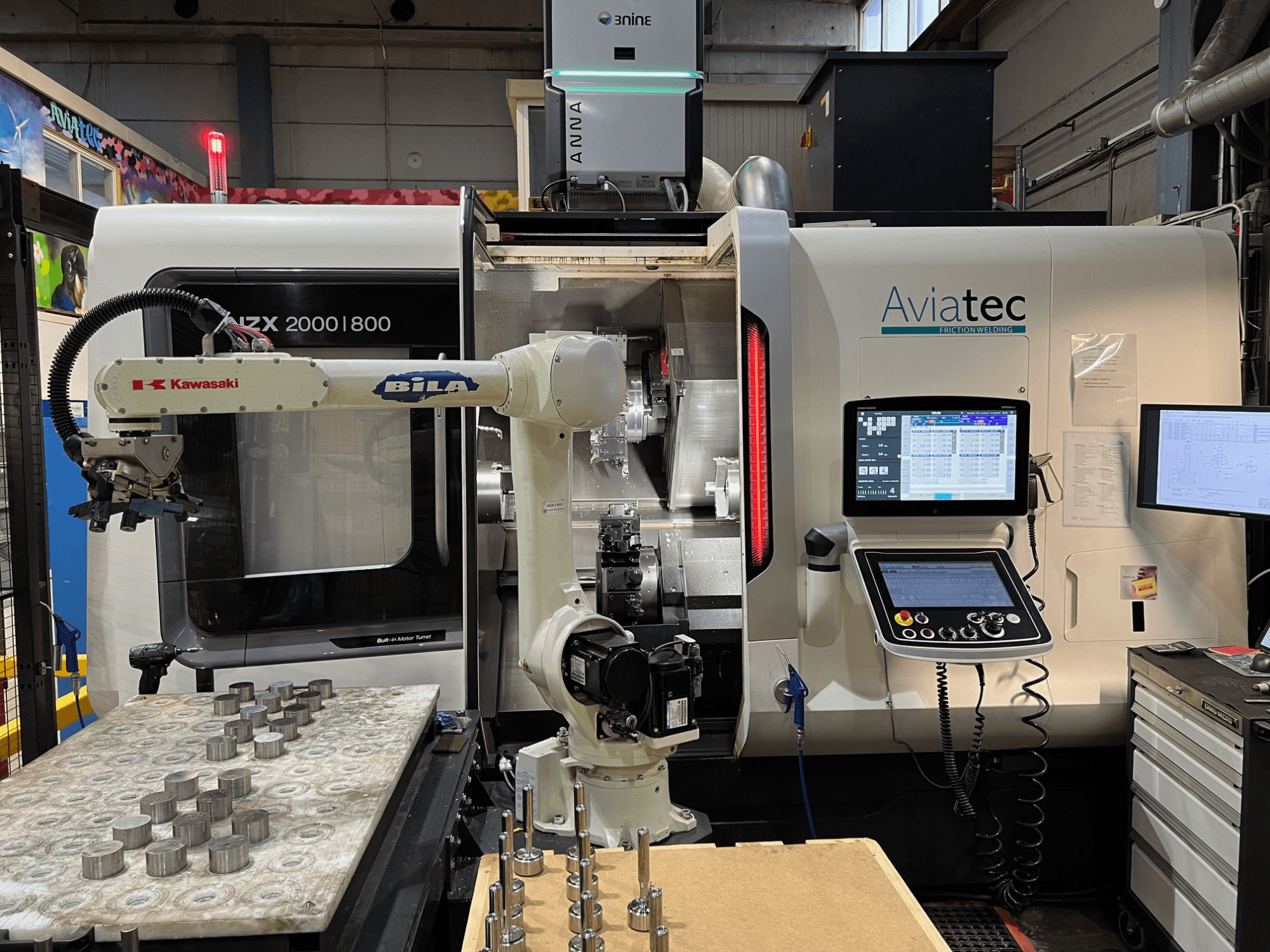 Horizontal turning machine DMG MORI NZX 2000/800SY with Kawasaki robotic arm, control panel, and metal components on worktable.