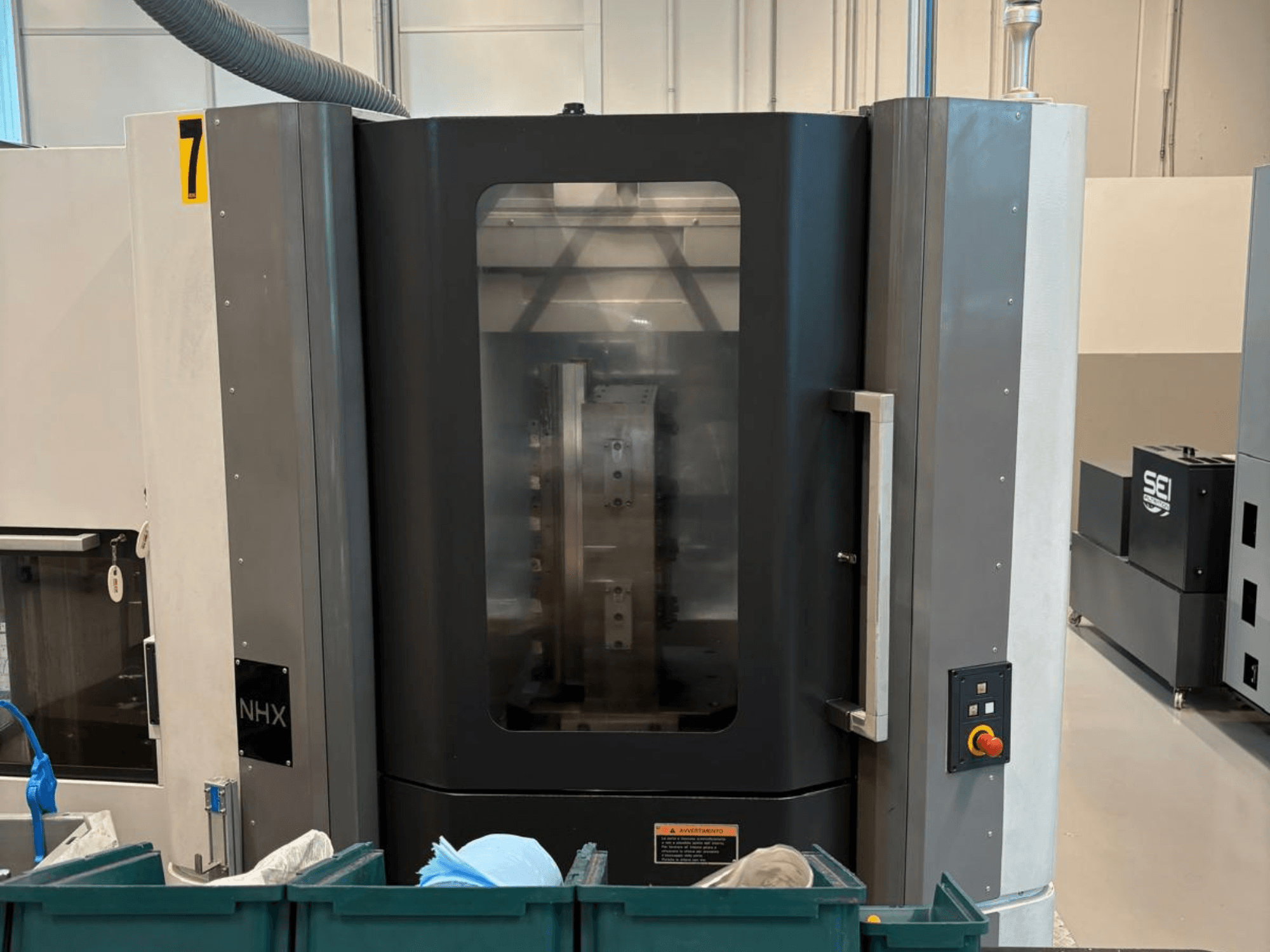 NHX CNC machine in front view, showing control panel and clear display; located in a workshop with green bins nearby.