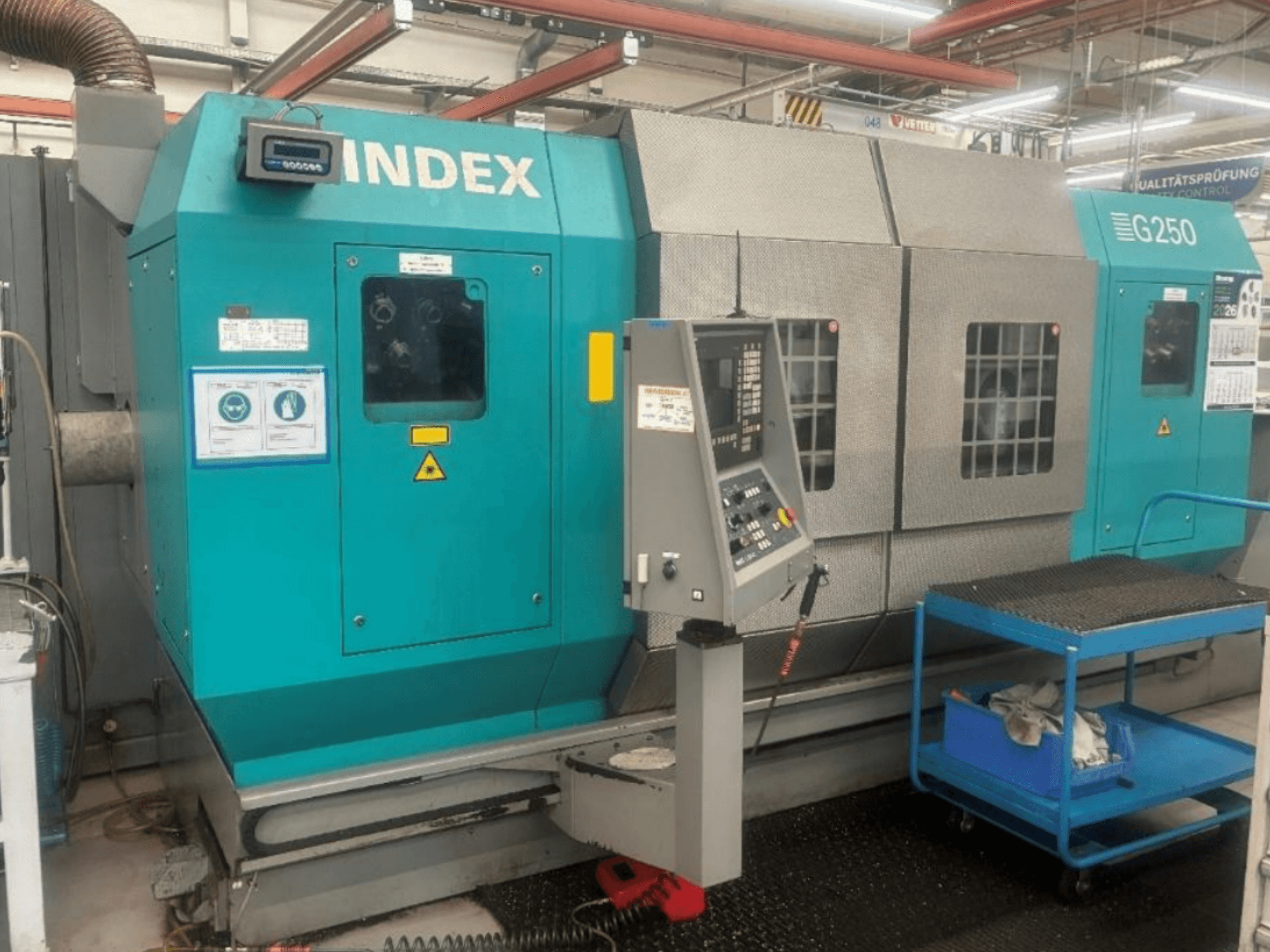Horizontal turning machine INDEX G250, front view showing control panel and metallic housing, with safety labels and a blue cart.