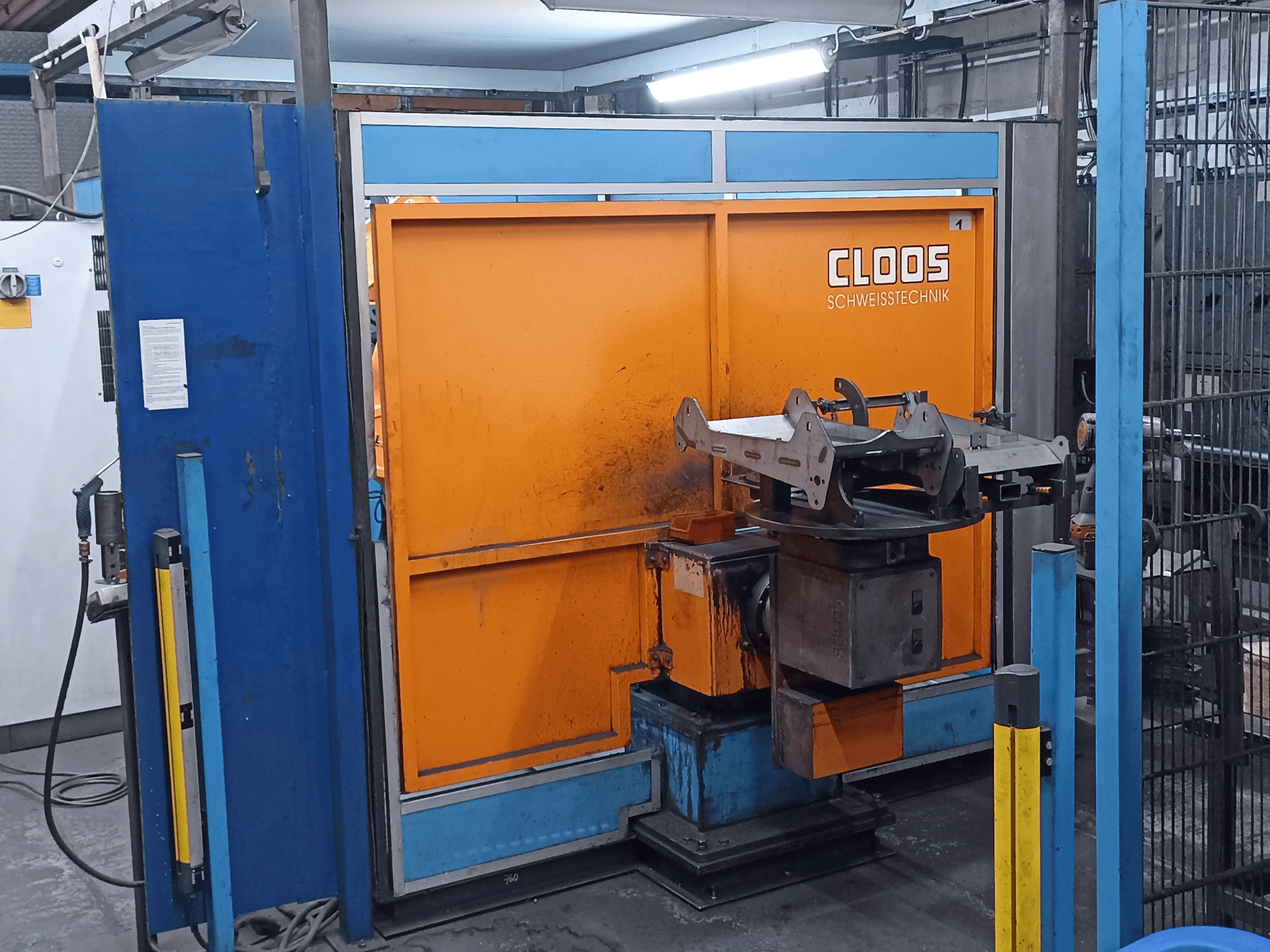 CLOOS Romat 320 welding robot in a workshop, front view, with an orange control panel and metal workpiece on a rotating base.