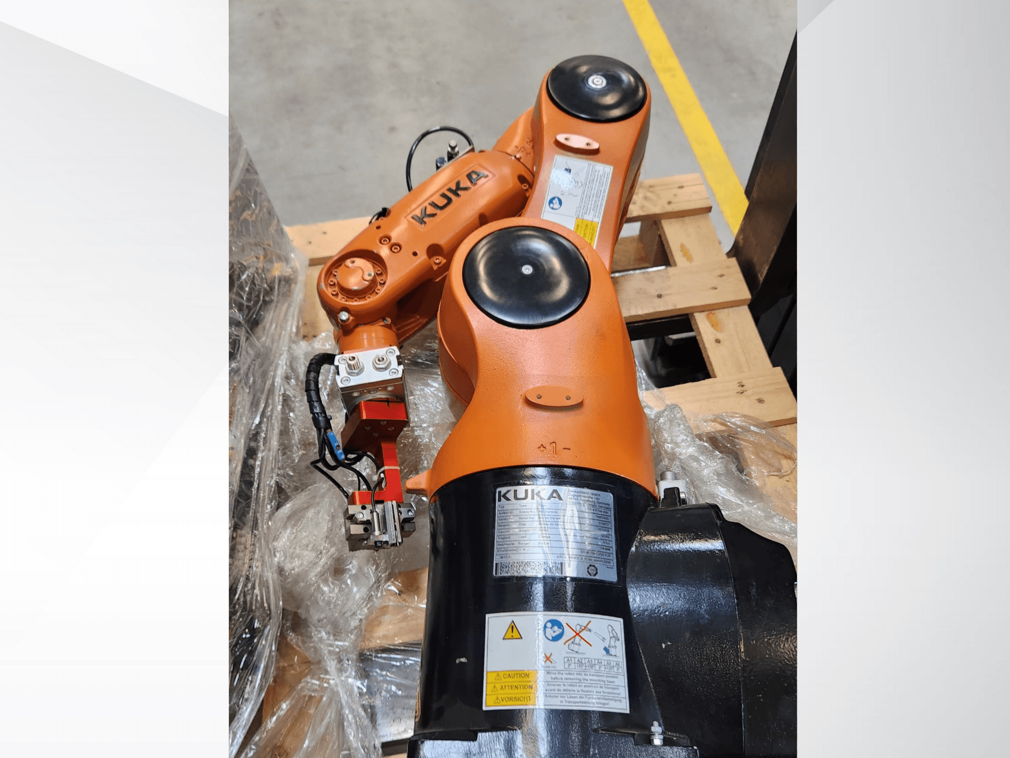 KUKA robotic arm with orange finish, side view, featuring a gripper and control wiring on a wooden pallet.