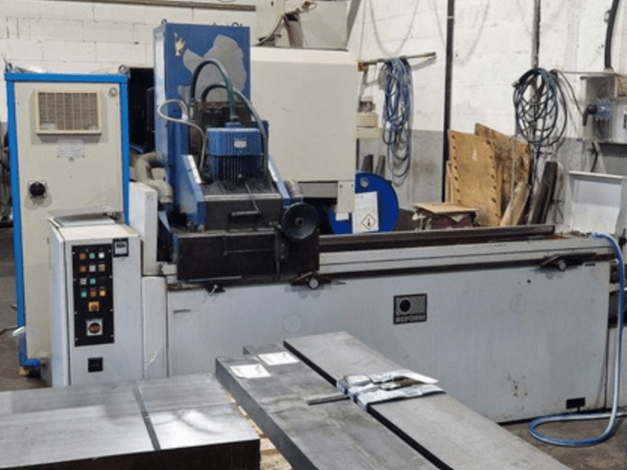 Reform Surface Grinder in front view, showing control panel, grinding area, and metal workpieces on table.