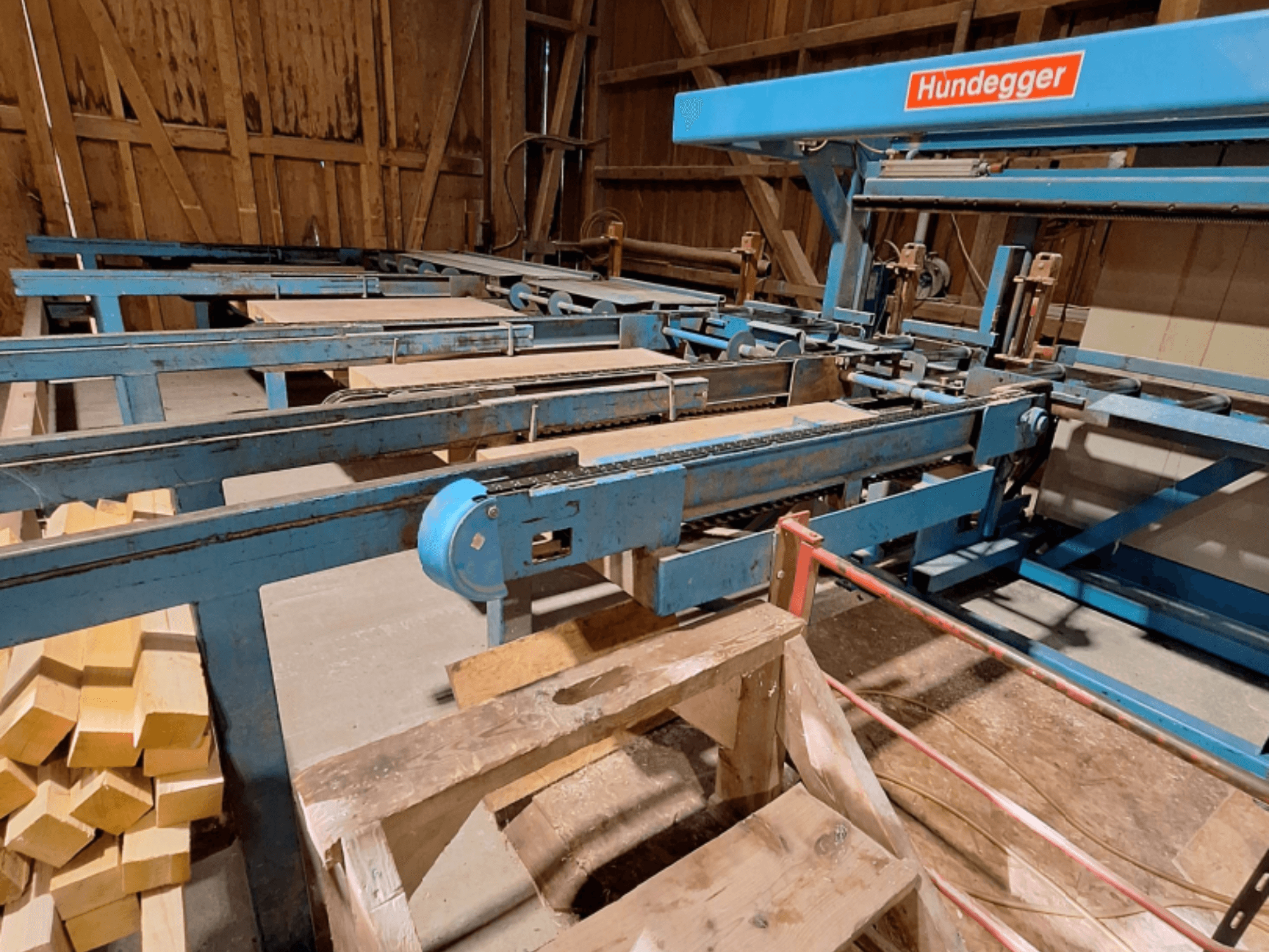 CNC Wood Machining Centre HUNDEGGER K2-A in a barn, featuring blue metal frame, conveyor system, and stacked wooden beams.