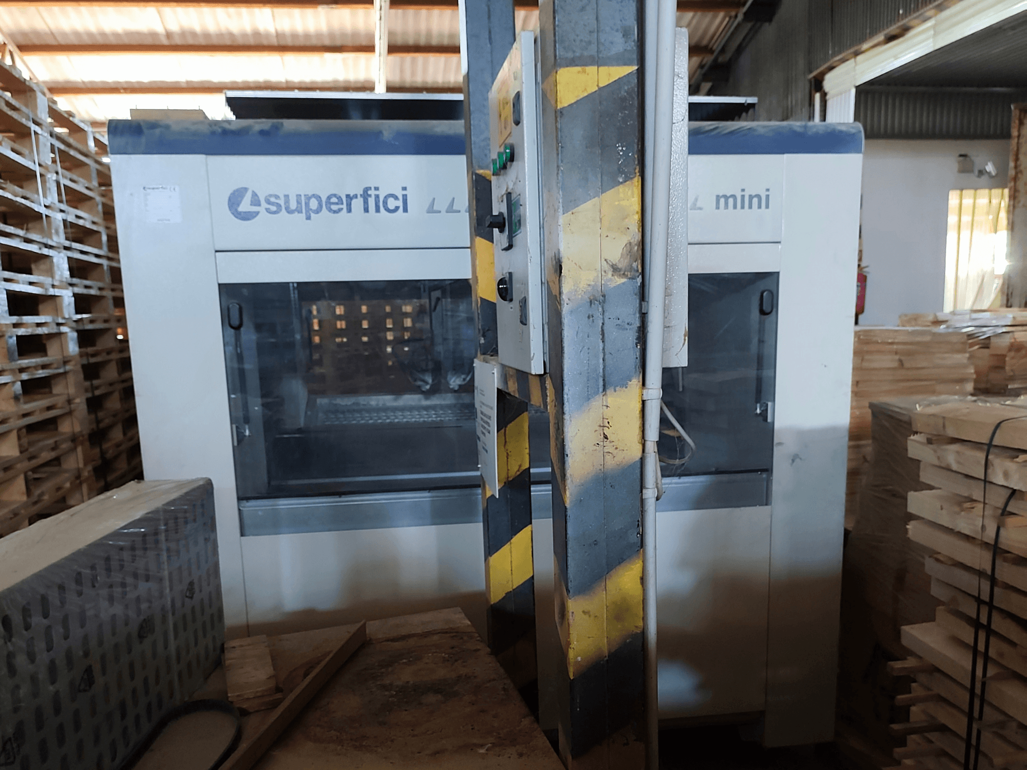 SCM Superfici Mini wood finishing machine in a front view, showing control panel and surrounding wooden pallets.