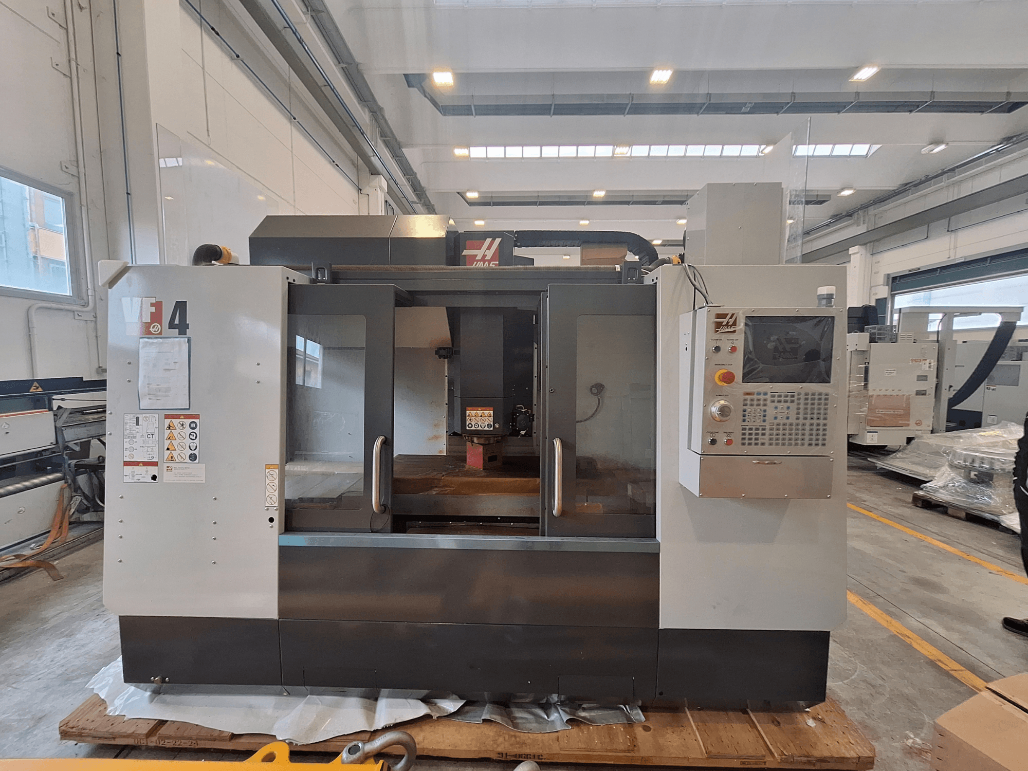 HAAS VF-4 vertical machining center, front view, showcasing control panel, work area, and safety labels in a warehouse setting.