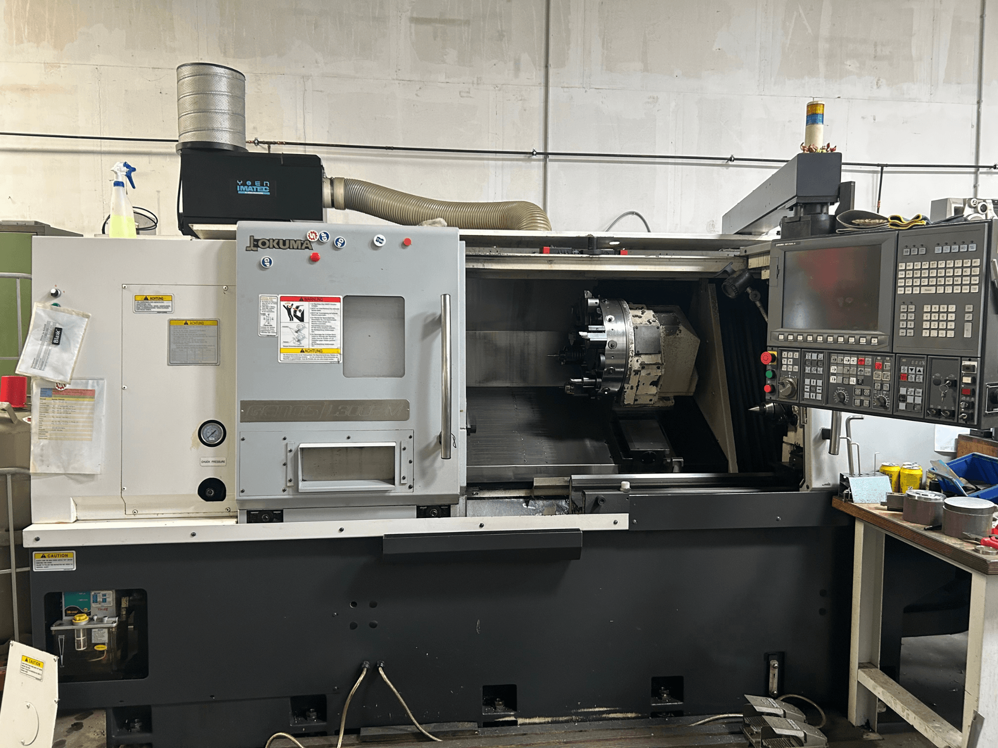 Front view of an Okuma Genos L300E-M horizontal turning machine with control panel, visible spindle and various setup tools.
