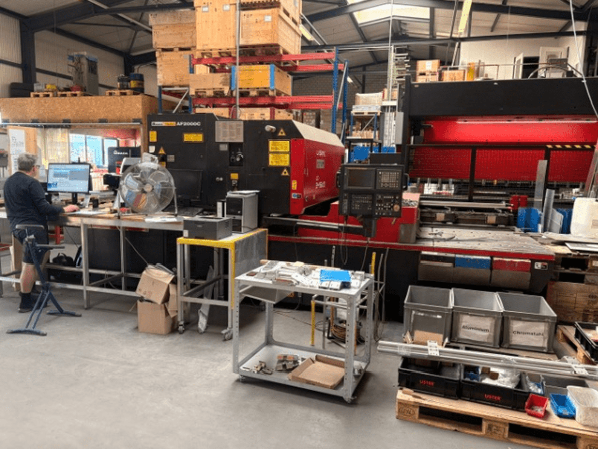 AMADA LC-2415αIII CO2 laser cutting machine in a workshop setting with a computer workstation visible in the foreground.