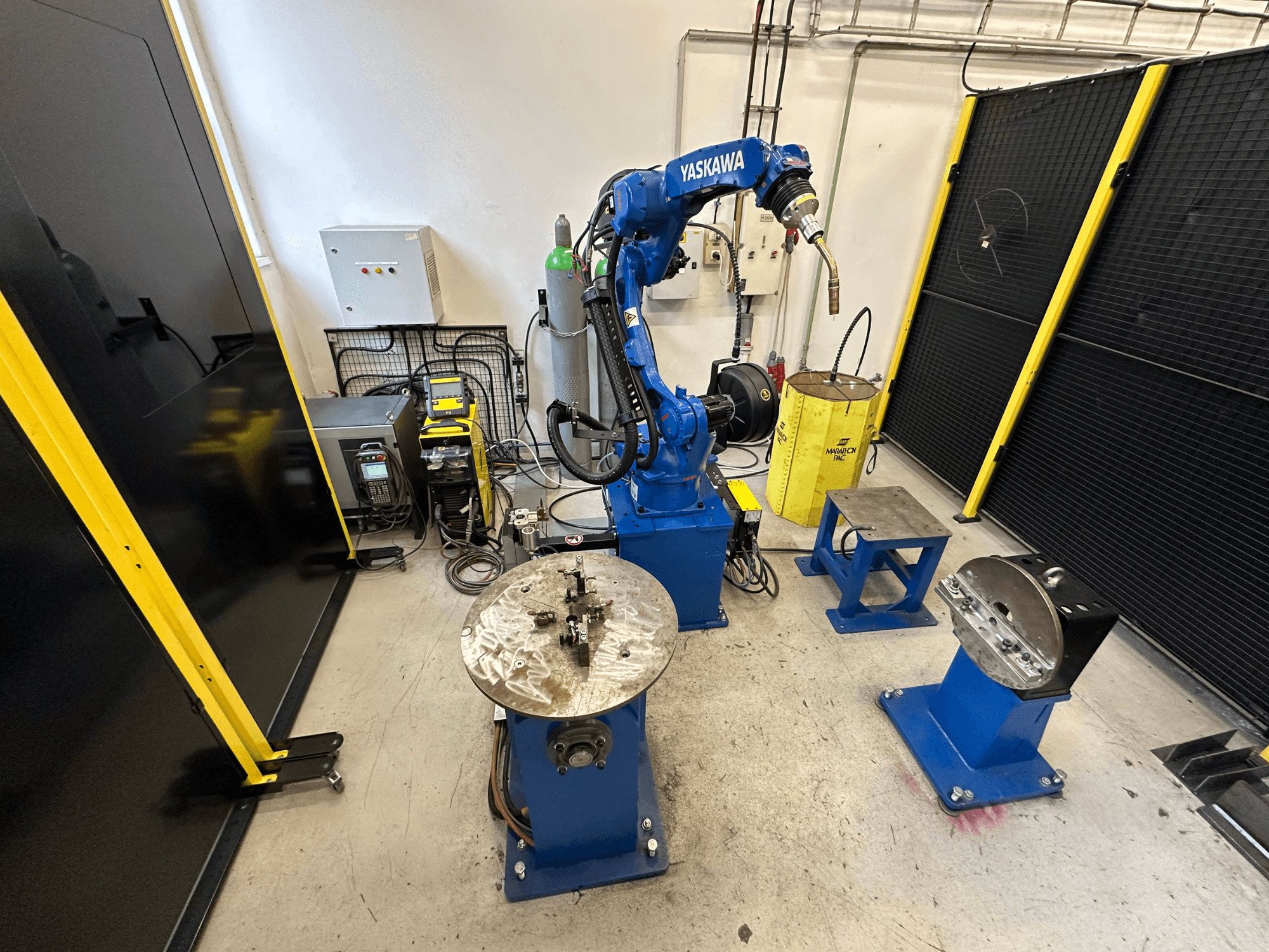 Yaskawa robotic arm in blue with two work tables, welding equipment, and safety barriers; industrial workspace front view.