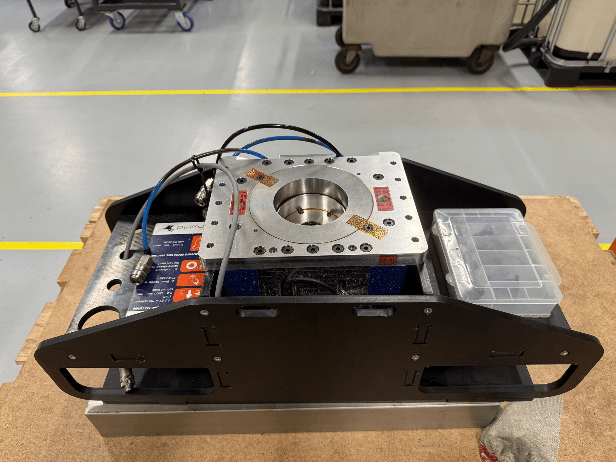 Machined metal component with blue cables, accessories case, and flat base, shown from top view, ideal for mechanical applications.