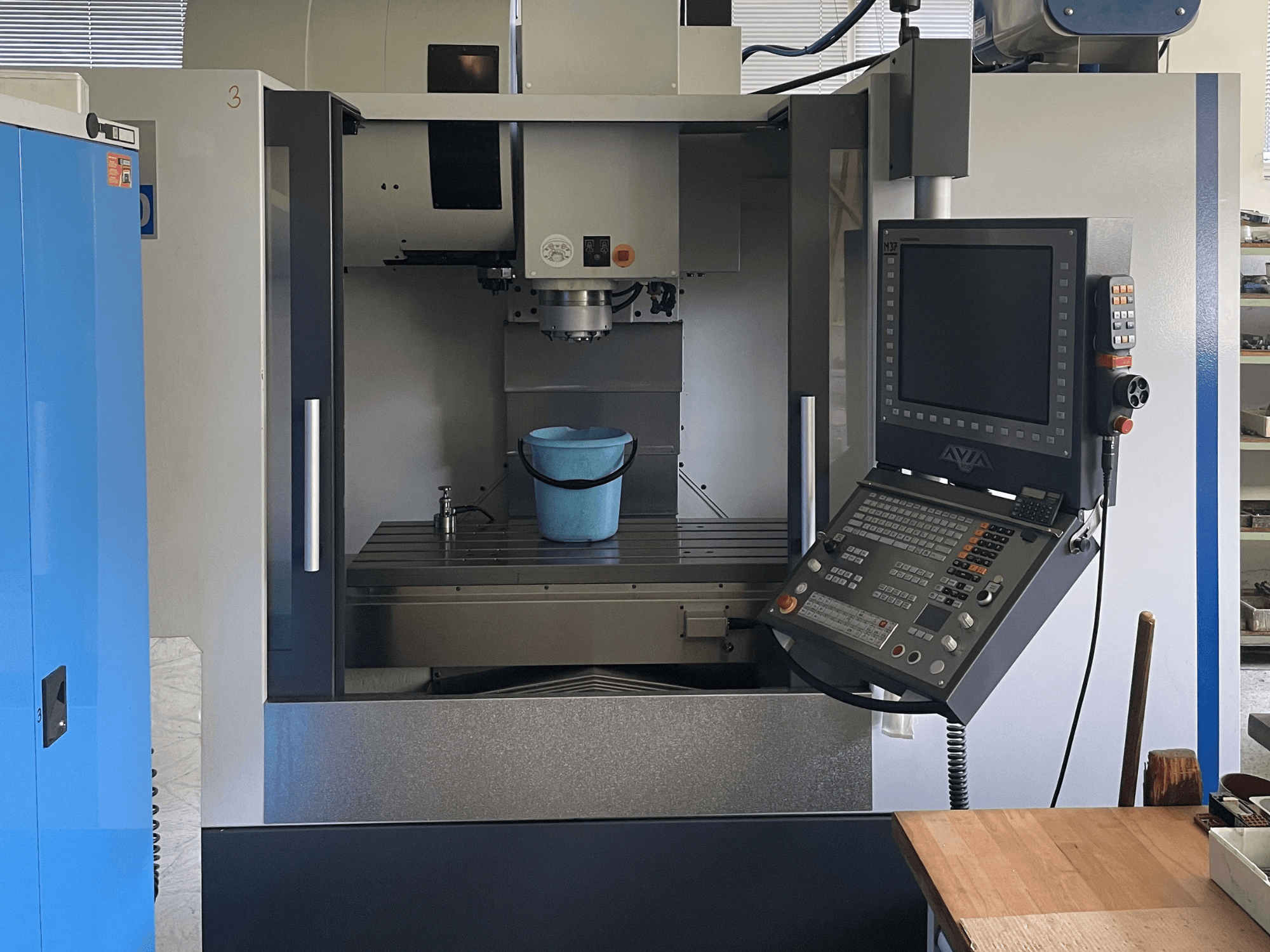 Avia VMC 1000 vertical machining centre in front view, featuring a blue bucket on the worktable and a control panel.