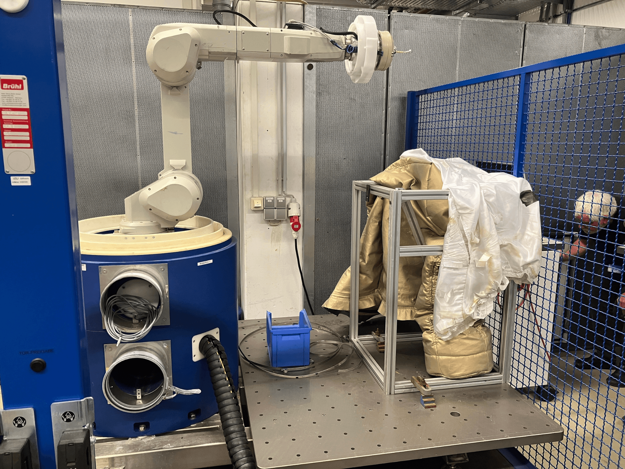 Robotic arm near a blue chamber, holding a white and gold suit on a metallic frame, side view with safety mesh background.