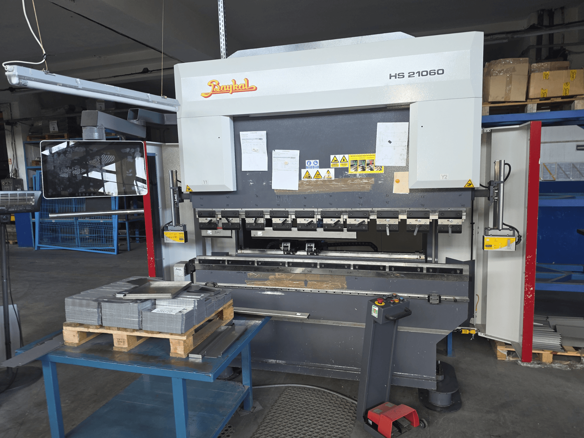 Baykal APHS 21060 press brake in a workshop, side view showing control panel, stacked metal sheets, and safety signage.