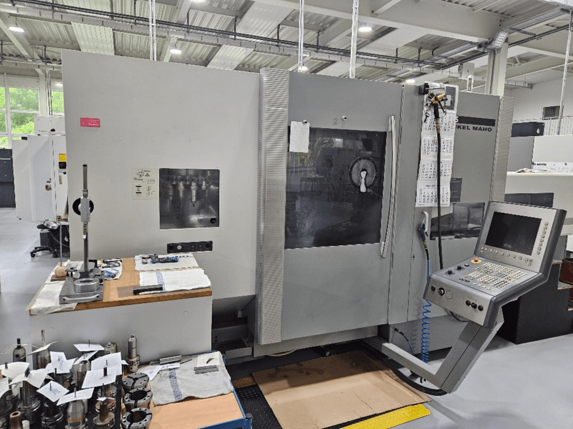 A front view of a DMG Mori DMU 70 machine with control panel, tools on a wooden table, and various workpieces nearby.