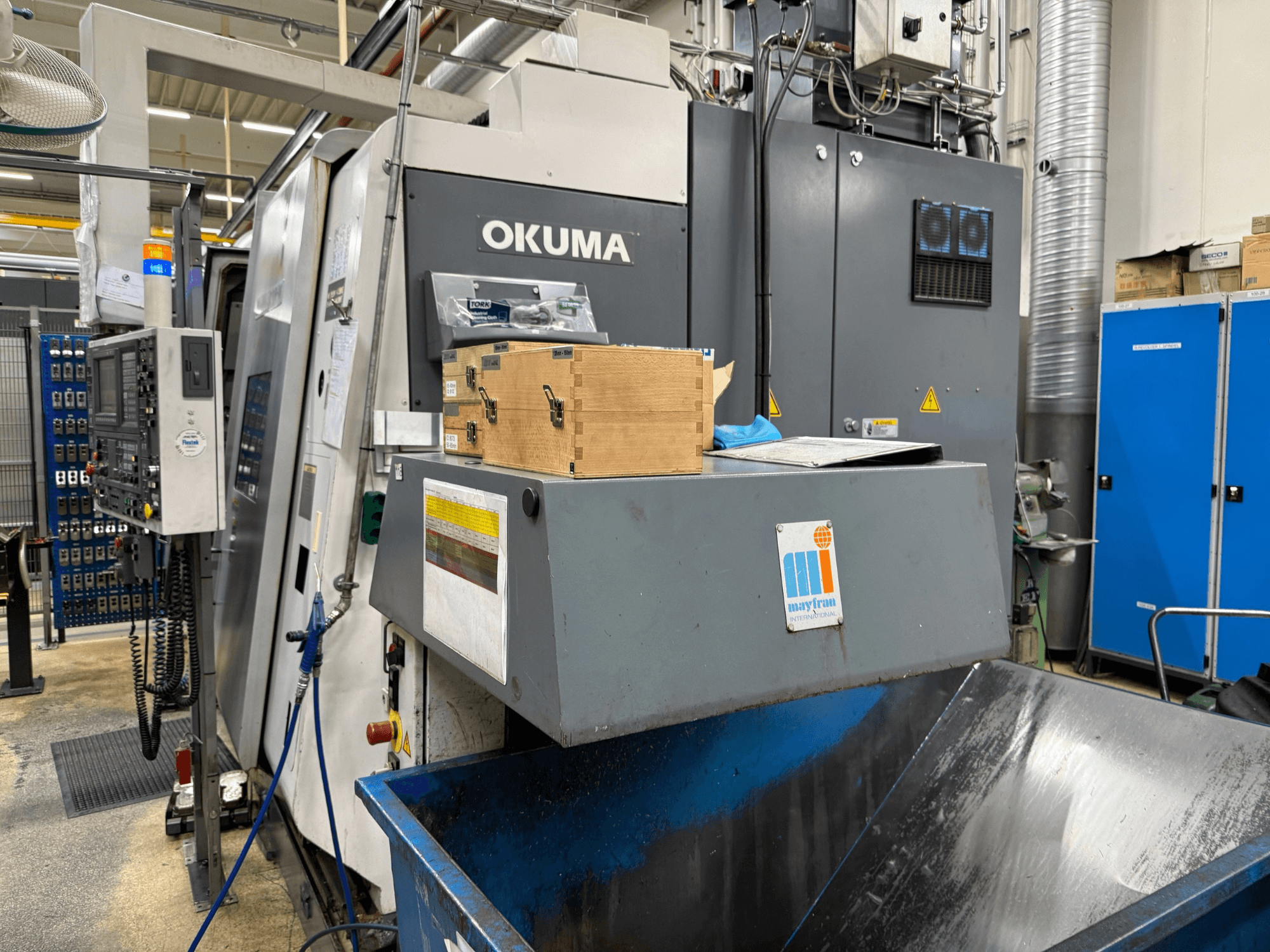 Okuma CNC machine in factory, front view; features control panel, wood boxes, and blue waste bin with Mayfran logo.