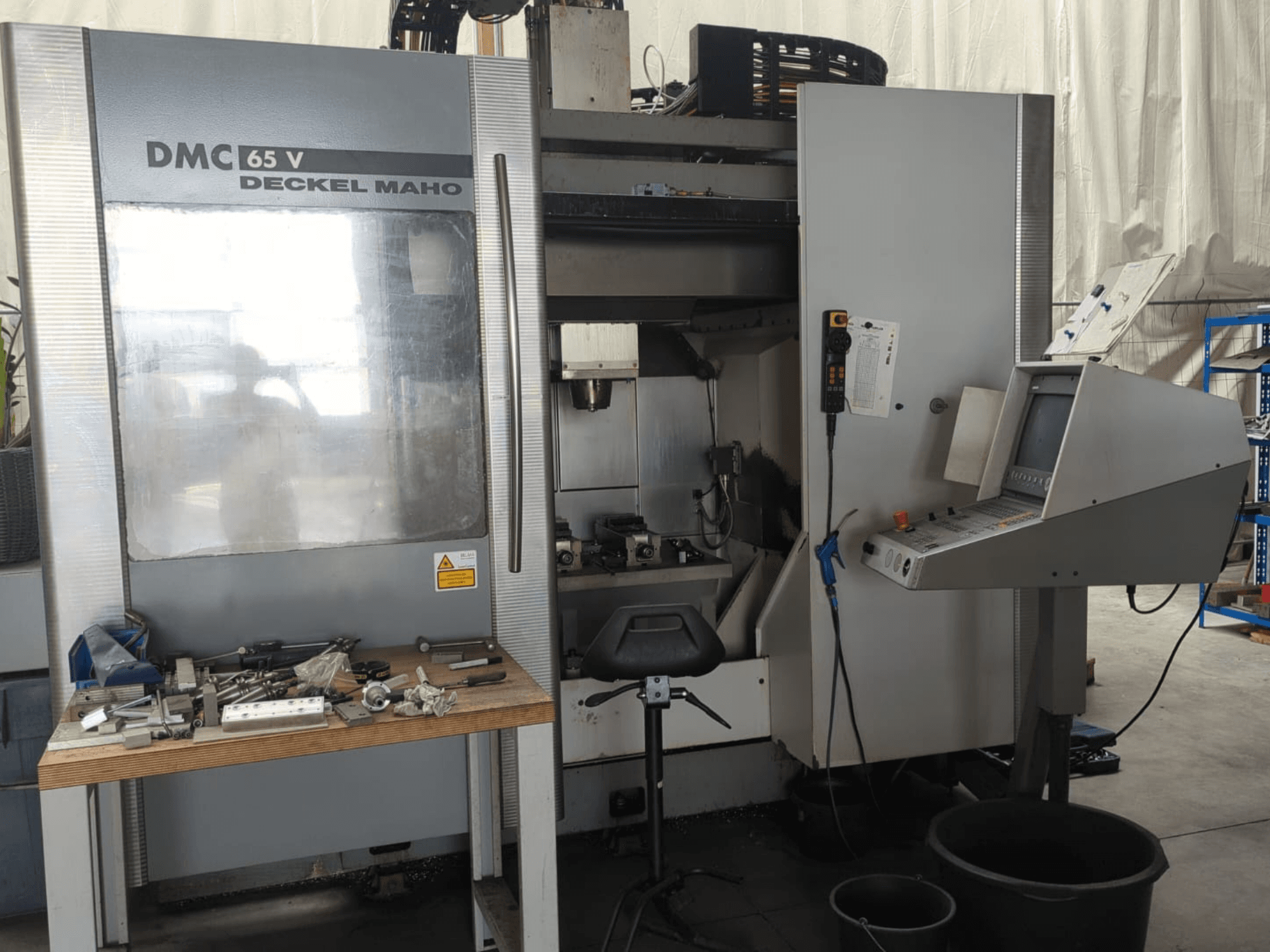 Front view of a DECKEL MAHO DMC 65 V vertical machining centre with control panel, tools on a table, and workspace setup.
