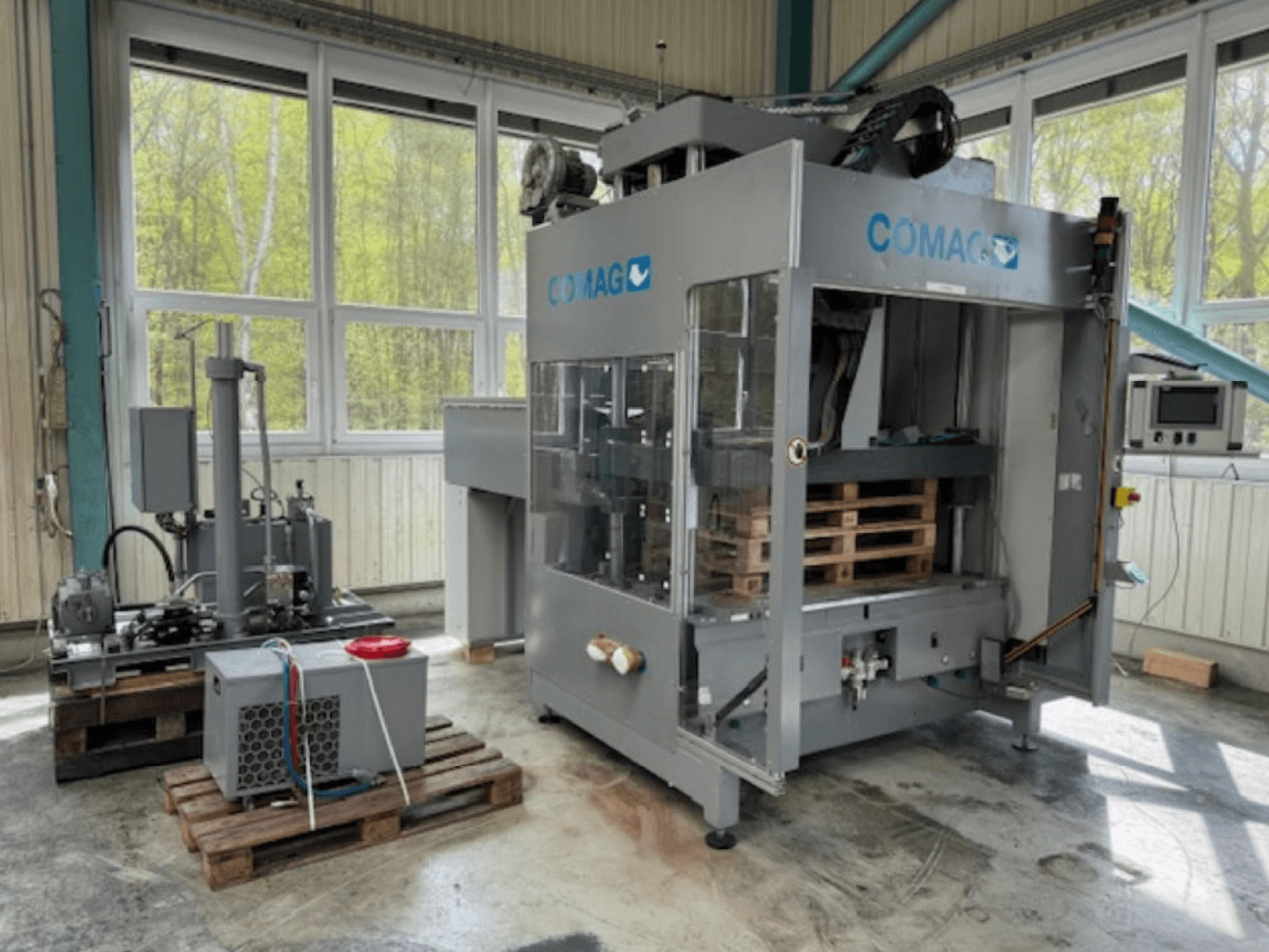 COMAG pallet wrapping machine in industrial setting, front view; features pallets and hydraulic components visible.