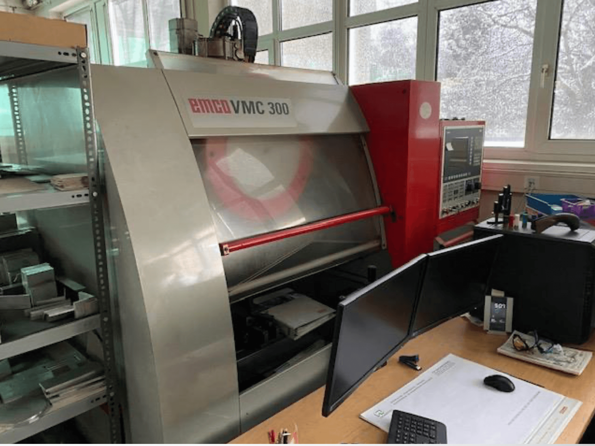 EMCO VMC 300 vertical machining centre in a workshop, side view showing control panel and nearby workstation with dual monitors.