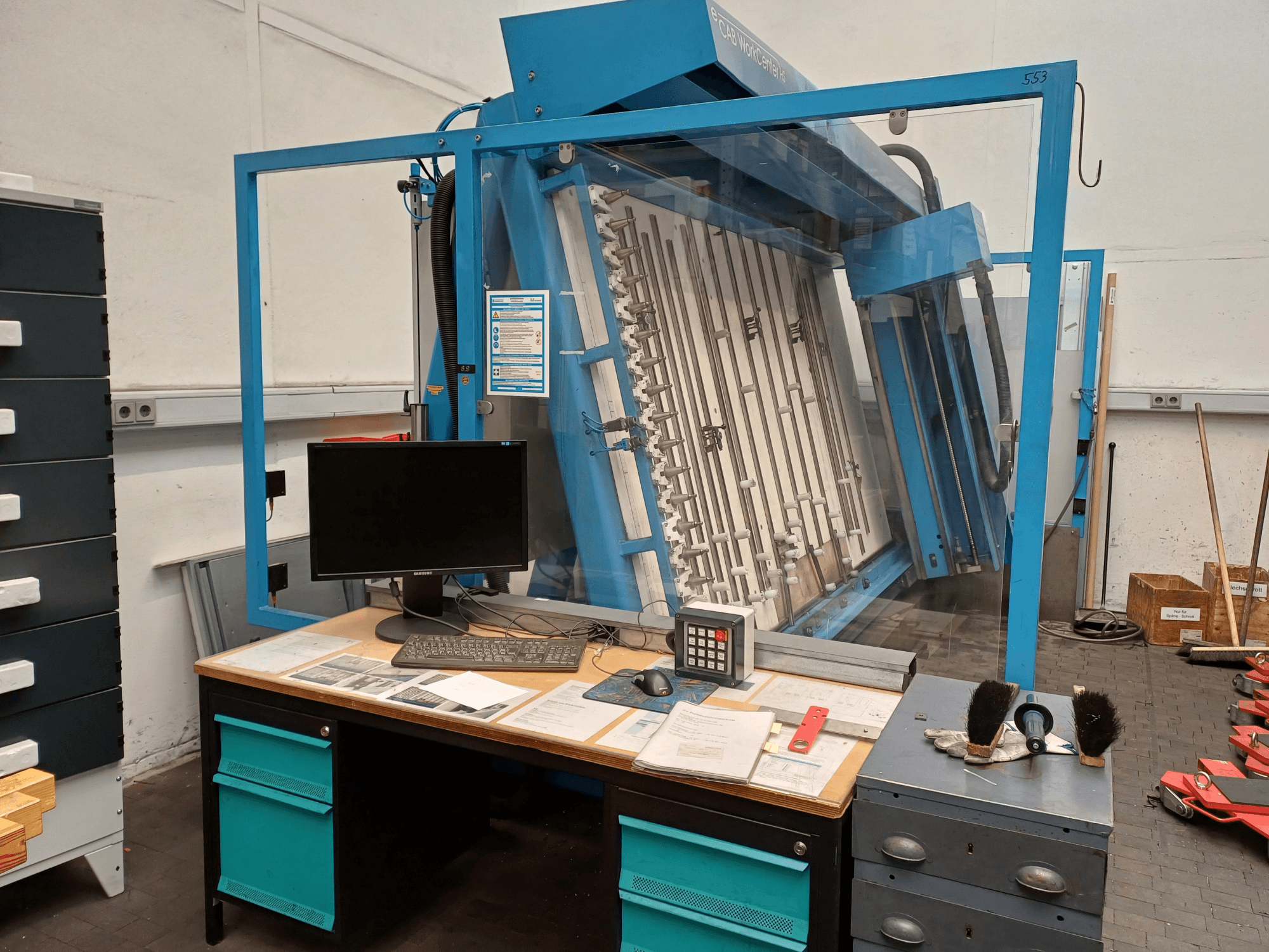 Blue glass display case with machinery inside, a monitor, and tools on a desk, photographed from the left side view.