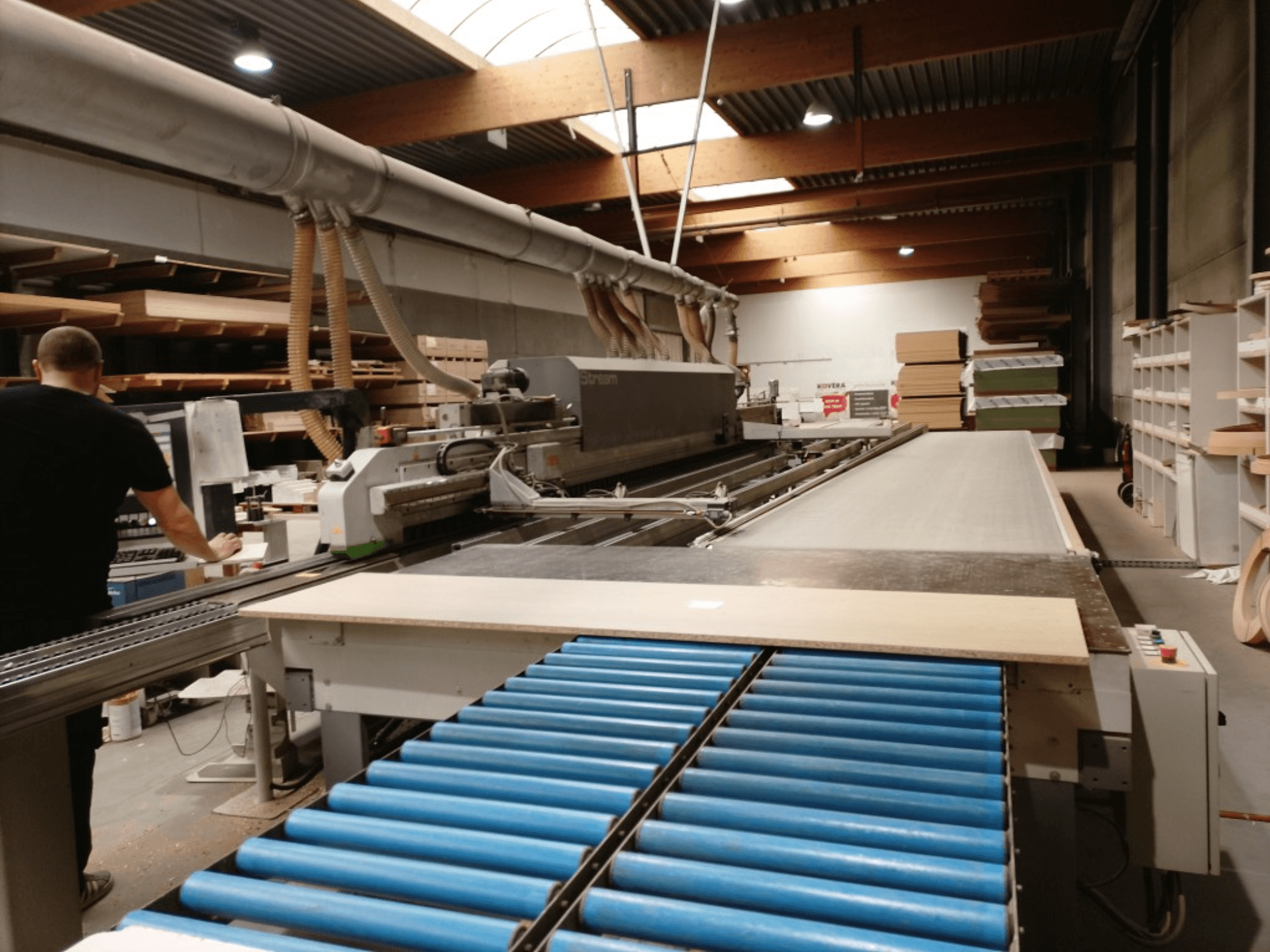 BIESSE Edge Stream B1 edgebander, side view showing operator, blue rollers, and wood panel on conveyor.