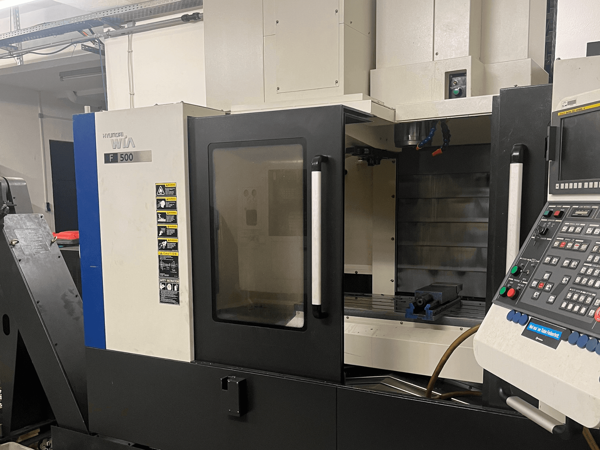 Hyundai Wia F500 vertical machining center, front view, showing control panel, work area with a blue vise, and warning labels.