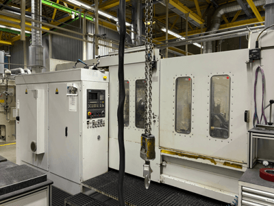 Industrial CNC machine with control panel, side view; features windows, chains, and metal components in a factory setting.